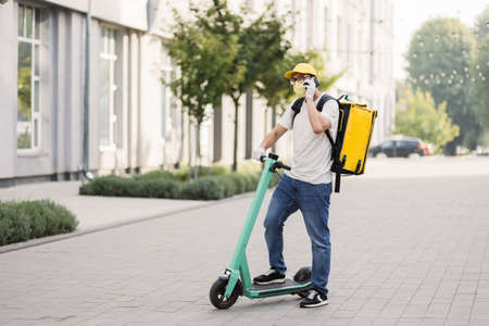 Side view of delivery man in face mask using smartphone and riding push scooter in the city. Young delivery man with thermal backpack using smartphone while riding scooter during covid pandemic.の写真素材