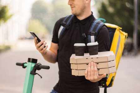 Close up of young delivery man calling by mobile phone to client outdoor. Courier delivering food and hot drinks outside. Guy using smartphone at city street. Courier with pizza and coffee.の写真素材