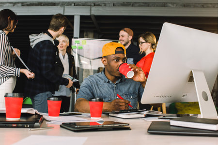 Cheerful African man drinking coffee and working on computer while sitting at the table in modern office place. His young coworkers standing and talking on the backgroundの写真素材