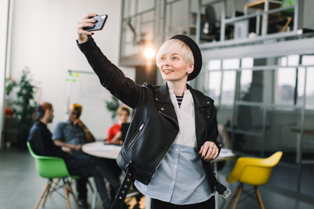 Cheerful girl in black hat and leather jacket making video call via smartphone. Young woman waving hand and smiling at phone screen. Video call concept.の写真素材