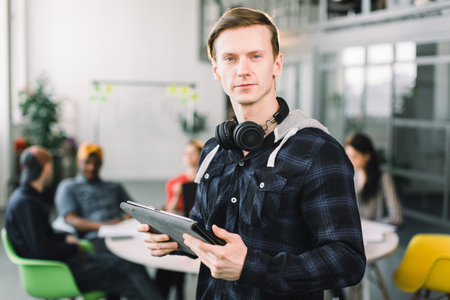 Half-length portrait of cheerful young manager or designer dressed in casual outfit and earphones, having work break in office, holding digital tablet. successful designers team having productive job.の写真素材