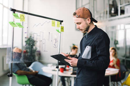 Young man in casual outfit and cap working with datas and diagrams on glass office wall with sticky notes, while his coworkers discussing ideas and brainstorming.の写真素材
