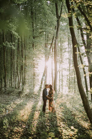 Sunset in the forest. Young Caucasian couple, pregnant woman and handsome man, standing face to face, walking in the forest, on the background of beautiful sunset in summerの写真素材