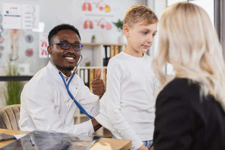 Healthcare and pediatry concept. Smiling confident African male gp doctor checking teen boys lungs and breathing during his regular scheduled visit to family clinic with motherの写真素材