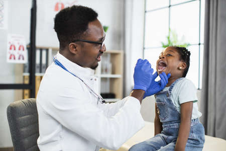 Lovely little afro-american child girl sitting on the couch at doctors office, while handsome black male doctor examining her throat and mouth with spatula. Pediatrics conceptの写真素材