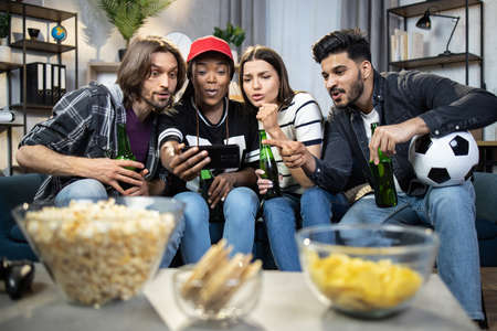 Four young people watching soccer match on modern smartphone while sitting together on couch. Multiracial friends emotionally supporting favorite team while staying at home.の写真素材