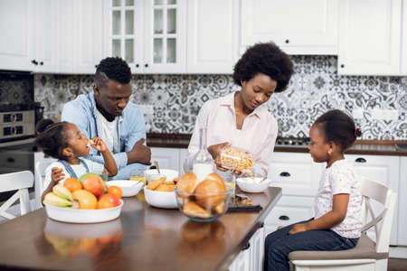 Positive african parents giving cereals with milk to their daughters for breakfast. Happy family of four enjoying tasty breakfast on modern kitchen.の写真素材