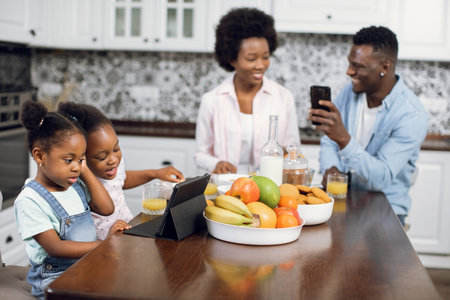 African american parents sitting on kitchen and using smartphone while their cute daughters watching...