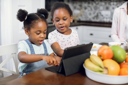 Pretty african american girls watching cartoons on digital tablet while sitting together on bright kitchen. Two sisters using modern gadgets during breakfast at home. Children and technologies conceptの写真素材