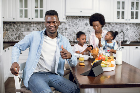 African man smiling, gesturing like and looking at camera while working on digital tablet on...