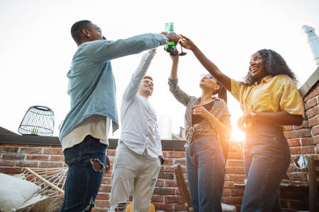 Cheerful young men and women in casual wear toasting with alcoholic cocktails during party. Multicultural friends having fun together on rooftop.の写真素材