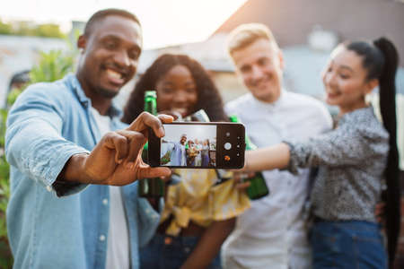 Four multicultural people in stylish clothes using modern smartphone for taking selfie during roof party. Happy young friends smiling, drinking, chatting.の写真素材