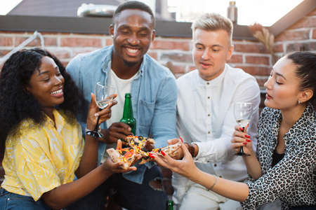 Smiling young men and women doing cheers with pieces of delicious pizza on roof top. Multicultural friends spending leisure time with fun. Weekends for party.の写真素材