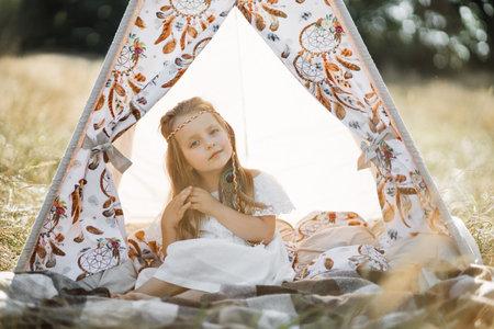 Adorable little blond girl sitting in boho teepee tent. Cute girl child, wearing white dress and...