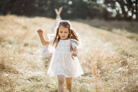 Cute girl is running and having fun in a summer field on sunset. Child pretends to be native american, is in white dress and has feathers in the hair. Stylish blurred wigwam on the backgroundの写真素材
