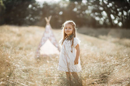 Summer sunshine in the field. Childhood concept. Portrait of confident cute little native american girl in white dress and feather accessories, posing in the wild field. Wigwam on the backgroundの写真素材