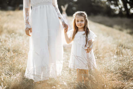 Close up of happy little child girl in white dress, holds hand of her mother and walks together in the summer field. Mother and daughter in boho dress spending time outdoorsの写真素材