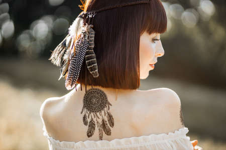 Close up rear outdoor portrait of attractive hippie boho indian american woman wearing brown feathers in short dark hair, and white blouse with bare shoulders. Sleep catcher tattoo on the backの写真素材