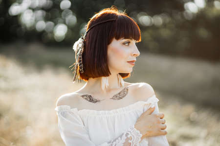 Wild nature, summer, ethnic tribal people. Profile of dreamy stunning young native american woman in white dress, with feathers in hair and body art tattoos, standing in the field and looking awayの写真素材