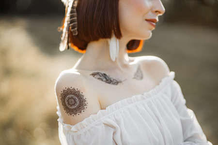 Close up native American woman neckline portrait. Cropped shot of pretty charming indian woman in white dress with bare shoulders, feather jewerly and body art tattoos, in the natureの写真素材