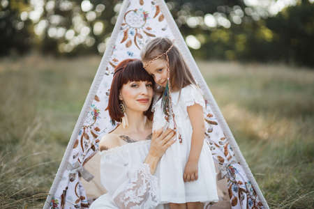 Portrait of happy native American mother and daughter at summer field. Woman and child wearing stylish clothes and feathers in the hair. Mom and daughter sitting near boho teepee tent in summerの写真素材
