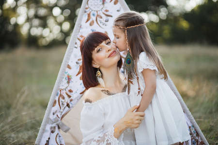 Outdoor shot of lovely Native American family near wigwam teepee in the field. Cute little girl with feathers in blond hair kisses her smiling cheerful motherの写真素材