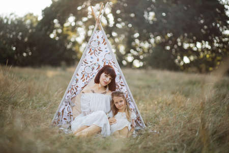 Front view of happy family mother and little girl, wearing stylish white boho dress and indian feathers in the hair, sitting in front of tee-pee tent at sunset in summer fieldの写真素材