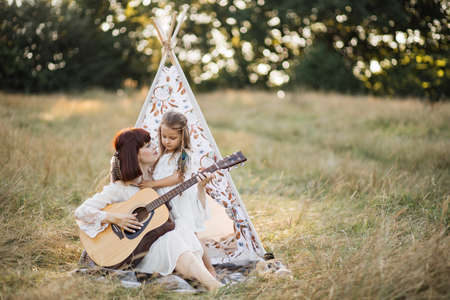Playing music and singing together. Smiling caring mother teaches her little daughter how to play the guitar, spending their leisure time outdoors in the field. Boho woman and little girl having funの写真素材