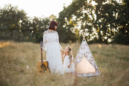 Happy woman in bohemian boho white dress, feathers and tattoos, walking with guitar on the field at sunset, together with her adorable little daughter. Teepee wigwam on the backgroundの写真素材