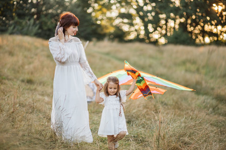 Happy family, holiday, mothers day concept. Charming mother and her cute adorable little daughter, launching a kite on nature at sunset. Boho hippie mom and child playing with kite in the fieldの写真素材