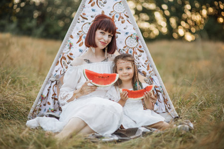 Close up of happy family, mother and daughter, sitting in front of boho wigwam teepee with fresh...