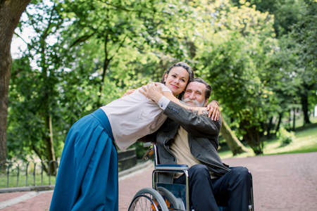 Disabled people, palliative care. Lifestyle summer portrait of senior bearded man in wheelchair hugging his young pretty hipster granddaughter, walking in park outdoors.の写真素材