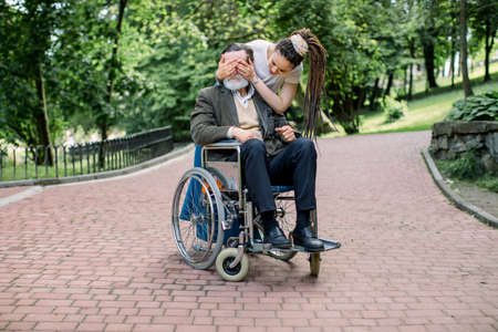 Handsome bearded senior man in wheelchair walking in a park. Young pretty granddughter or care giver is covering his grandfather or patient eyes doing a surprise.の写真素材