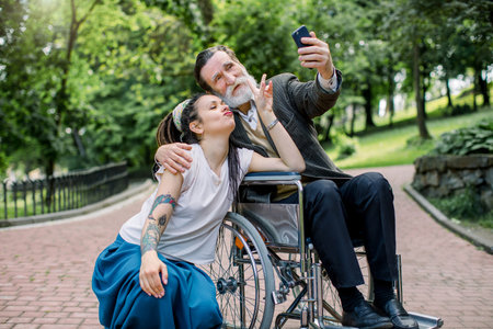 Hipster girl with long dreadlocks and handsome bearded senior man grandfather in wheelchair at park taking selfie. Lifestyle of disabled people. Care giver and patient on a walk.の写真素材