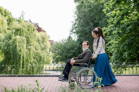 Side view of young modern hipster woman with long dreadlocks, assisting her disabled grandfather on wheelchair in park. Happy elderly man in wheelchair with young girl social worker in the parkの写真素材