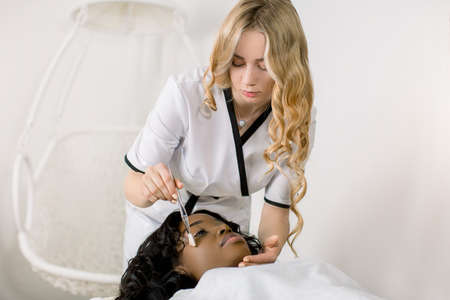 Young African woman having injection on forehead in salon, side view. Pretty young blond female doctor cosmetologist making facial treatment procedures using cosmetology brush and special oil cream.の写真素材