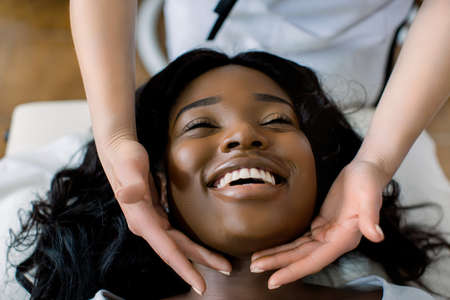 Close-up of hands of cosmetologist woman making special face massage to young pretty African smiling woman lying on couch in beauty salon. Concept of antiaging facial skin treatments and massage.の写真素材