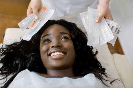 Aesthetic surgery. Close up of face of young African woman looking up and smiling while waiting for beauty injections in clinic, top view. Hands of female doctor cosmetologist holding set of syringes.の写真素材