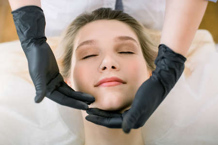 Close-up of hands of cosmetologist woman in black gloves making special face massage to young pretty Caucasian smiling woman lying on couch in beauty salon with eyes closed.の写真素材