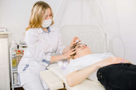 Indoor shot of pretty woman cosmetologist applying to her female client anti aging mask or cream. Young girl relaxing during moisturizing and rejuvenation procedures in beauty salon.の写真素材