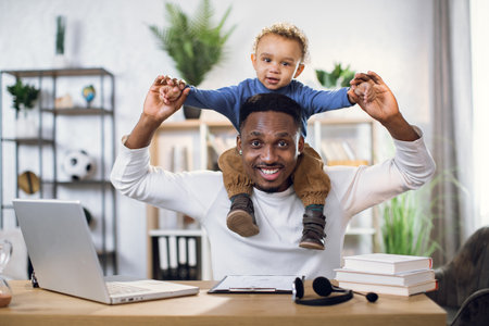 Young afro american father holding cute baby son on neck while sitting at table with portable laptop. Happy man and little boy smiling and looking at camera.の写真素材