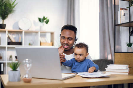 Afro american man sitting at table with cute baby boy and having video chat on laptop. Young father taking care of son and working on distance.の写真素材