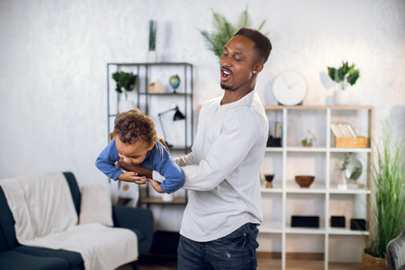 Cheerful african man in casual outfit spending free time with cute son and taking fun. Young father playing and carrying on hands baby boy at home.の写真素材