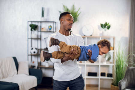 Cheerful african man in casual outfit spending free time with cute son and taking fun. Young father playing and carrying on hands baby boy at home.の写真素材