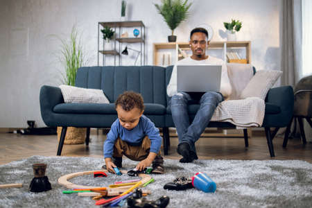 Focused male freelancer sitting on sofa and using modern laptop for remote work. Pretty boy playing with various toys while father working.の写真素材