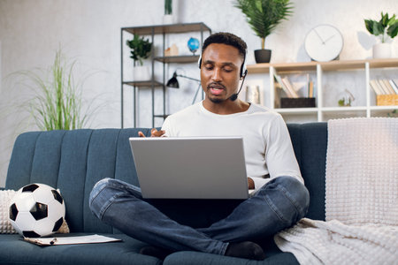 Casual african man in headset having video chat on modern laptop while sitting on cozy sofa. Young...