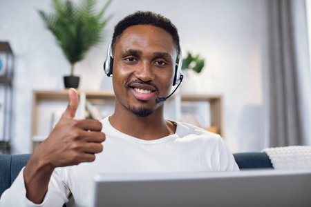 Smiling african man showing thumb up on camera while sitting on couch with headset and laptop. Happy...