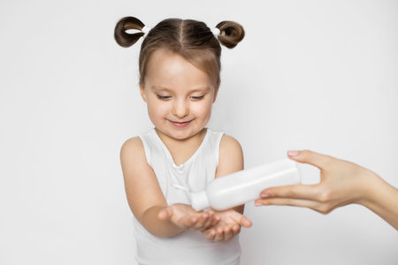 A little girl in white tank top and with two ponytails, standing on white background while hand of her mom pours a sanitizer on her hands. Sanitary precautions - hand disinfectionの写真素材
