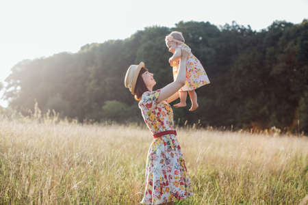 Happy smiling pretty mother wearing dress and straw hat, holding and raising up little baby daughter playing in beautiful summer fieldの写真素材