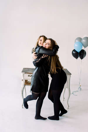 Two young pretty women in black dresses holding air balloons together, celebrating hen or birthday partystanding near the vintage white table with closets, isolated on white.の写真素材
